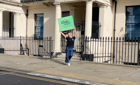 Esme holding a green Clubwell sign in front of a building with columns.